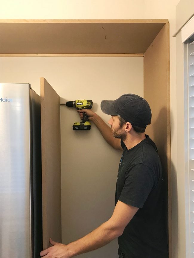John drilling vertical MDF panel into wood already placed near fridge in pantry